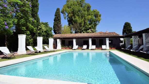 A rectangular swimming pool with clear blue water, surrounded by a tiled deck. There are white lounge chairs and a building with a tiled roof in the background. - Costa del Sol Villa Near Beach (San Pedro de Alcantara)