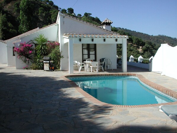 A pool area with a covered seating area, stone flooring, and a view of a hillside.