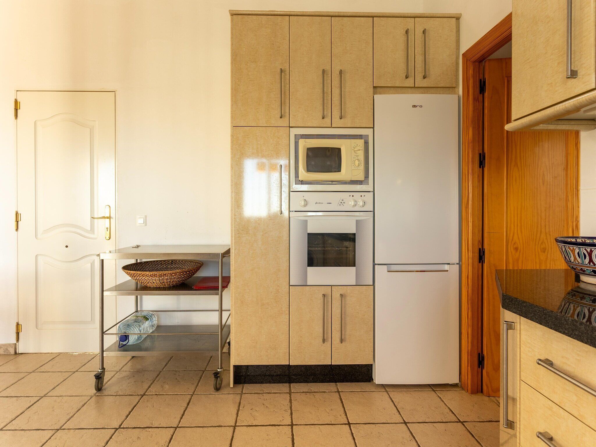 Une cuisine équipée d’un four à micro-ondes, d’un four traditionnel et d’un réfrigérateur, ainsi que d’un meuble en bois et d’un chariot en métal muni d’un panier.