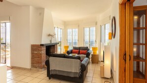 A spacious living room with a fireplace, a sofa, and a view of the sea through large windows. - Villa in Arenas with Sea Views and Private Pool (Arenas)
