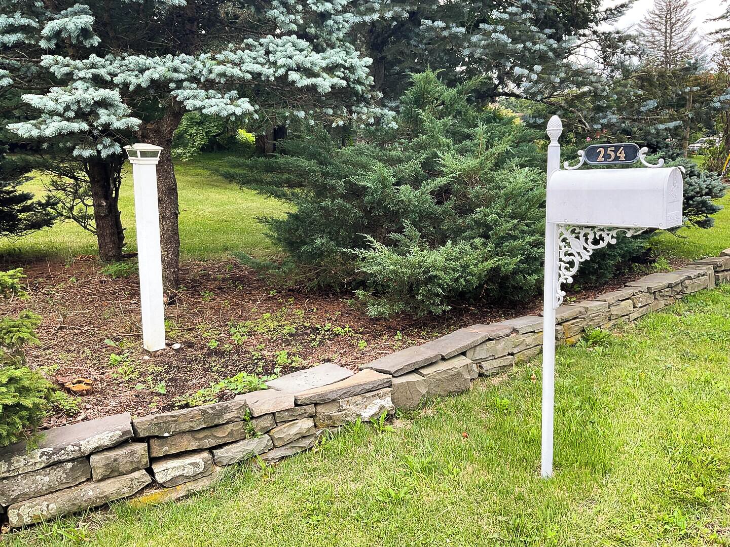 A mailbox with a number sign, a stone wall, and a tree in the background.