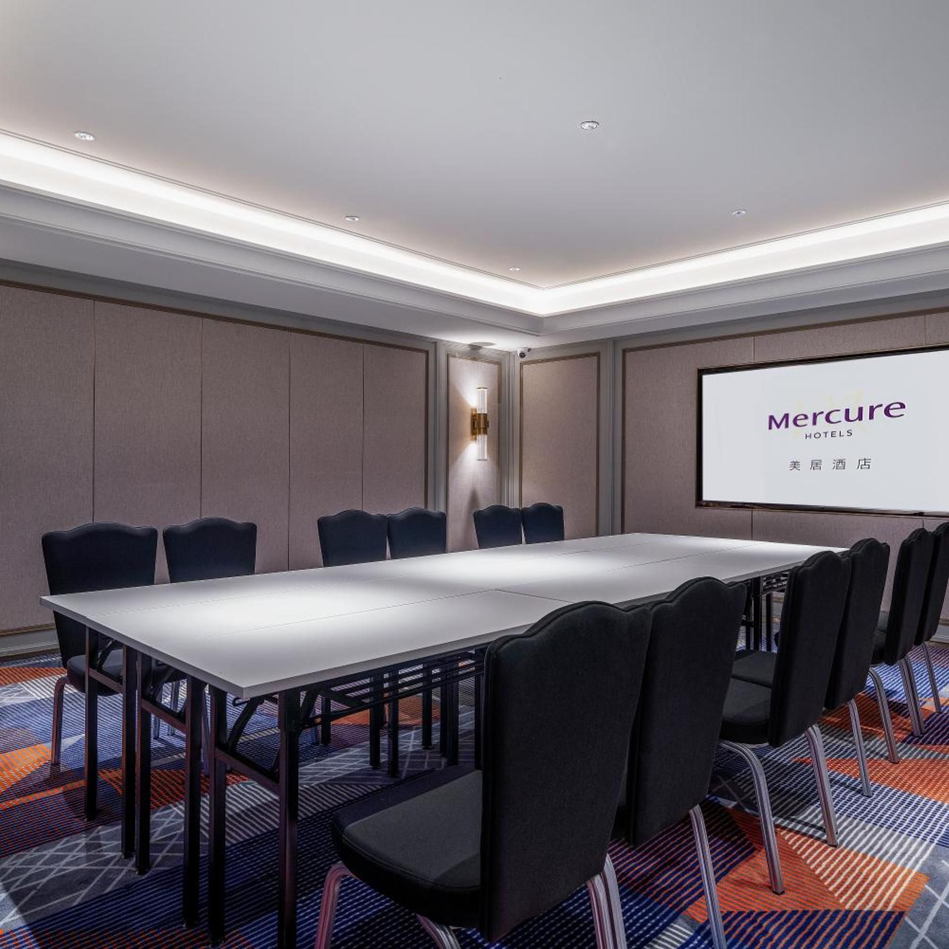 A conference room with a long table and chairs, a projector screen displaying 'Mercure Hotels', and a patterned carpet.