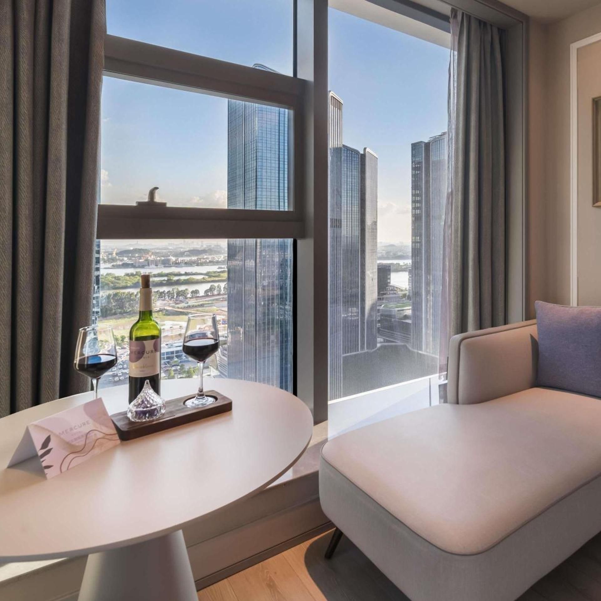 Quarto de hotel moderno com área de jantar, sofá e uma grande janela com vista para a cidade.