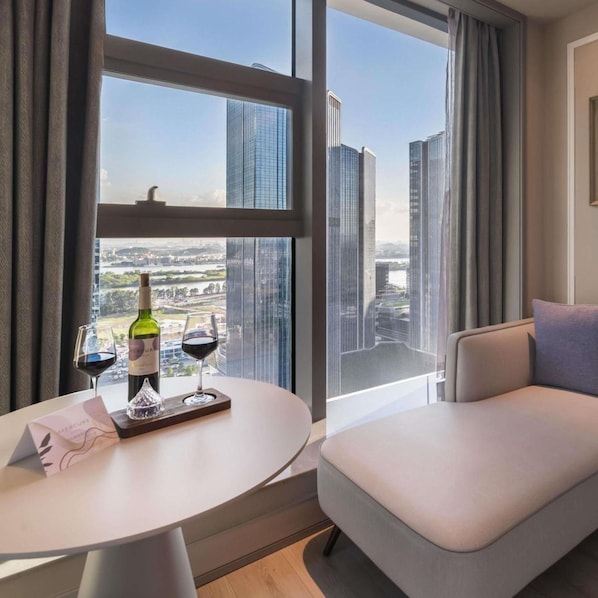 A modern hotel room with a dining area, a sofa, and a large window offering a city view.