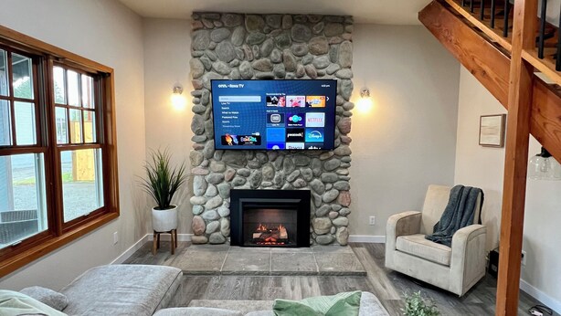 A living room with a stone fireplace, a flat-screen TV mounted above it, a wooden staircase, and a large window. - Forks Cedar House (Forks)