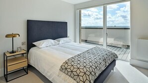 A modern bedroom with a large bed, a bedside table with a lamp, and a view of the sky through a sliding glass door. - Meadow Lane Retreat - City Center Gem (Hounslow)