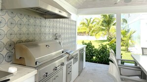 A modern kitchen with a built-in grill, white cabinetry, and a view of a garden with palm trees. - Treasure Cay, Palm Bay 7, Anchorage Drive (Treasure Cay)