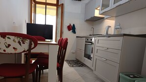 Une cuisine de taille réduite avec des meubles blancs, un four encastré et une fenêtre agrémentée d’un rideau.