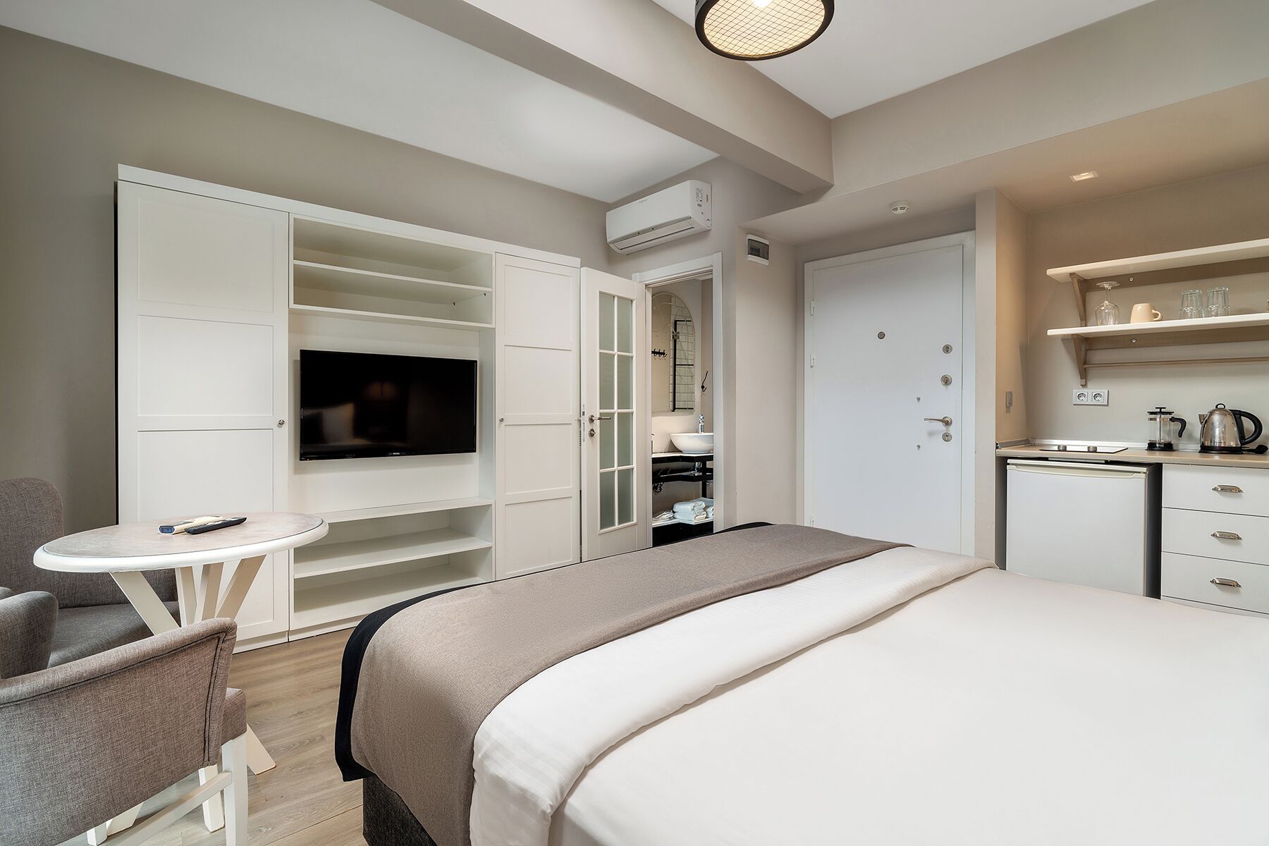 A modern hotel room with a bed, a TV, a small table, a chair, a kitchenette, and a wardrobe.