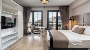 Habitación de hotel moderna con una cama grande, un televisor, una mesita con sillas y vistas a la ciudad a través de amplios ventanales.
