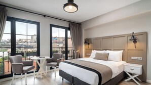 Habitación de hotel moderna con una cama grande, dos mesitas de noche, una silla y una pequeña mesa. Cuenta con ventanas amplias con cortinas, vistas a la ciudad y una lámpara de techo.