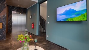 Un vestíbul amb sòl de marbre, una taula rodona amb un ram de flors, un televisor fixat que mostra un paisatge idíl·lic i una escala al fons.