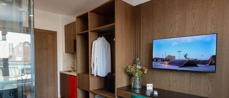 A modern hotel room with a wooden desk, a TV mounted on the wall, a bed with a blue blanket, and a small kitchenette area.