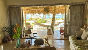 A spacious living area with a large window overlooking a beach, a ceiling fan, and a rustic wooden door. - Casa Taina- 5 Bungalows, swimming pool- OCEANFRONT w/Butler and Chef (Cabrera María Trinidad)