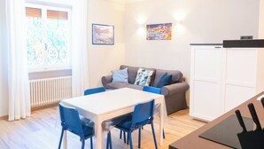 A compact living space with a dining table, blue chairs, a grey sofa with patterned cushions, a white cabinet, and two wall-mounted artworks. - Lemon Apartment with Garden and Sea View (Bordighera)