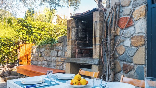 An outdoor dining area with a stone-built barbecue grill, a wooden table set for two, and a variety of fruits on the table. - Lemon Apartment with Garden and Sea View (Bordighera)
