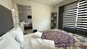 Un dormitorio con cama, armario, espejo, ventana con persianas y televisor en la pared.