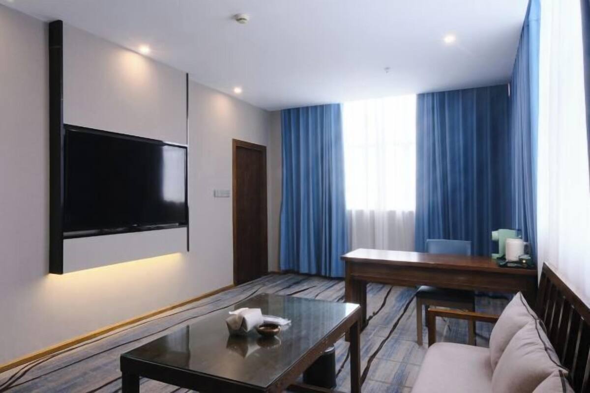 A hotel room with a flat-screen TV mounted on the wall, a wooden dining table, a sofa, and a large window with blue curtains.