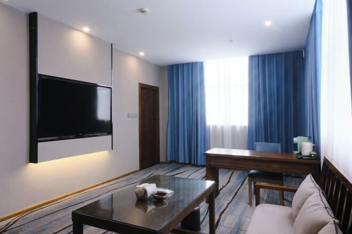 A hotel room with a flat-screen TV mounted on the wall, a wooden dining table, a sofa, and a large window with blue curtains.
