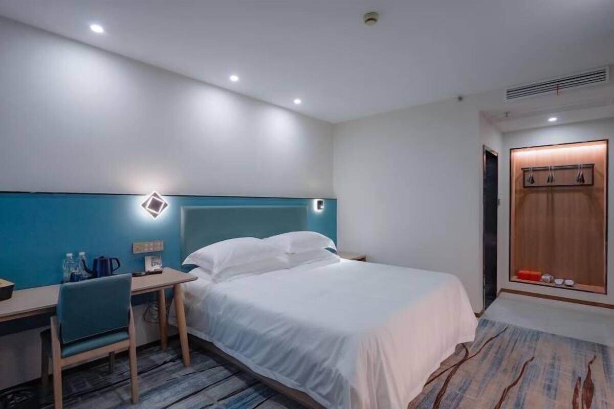 A hotel room with a bed, a desk, a chair, a wardrobe, and a small table with a lamp.