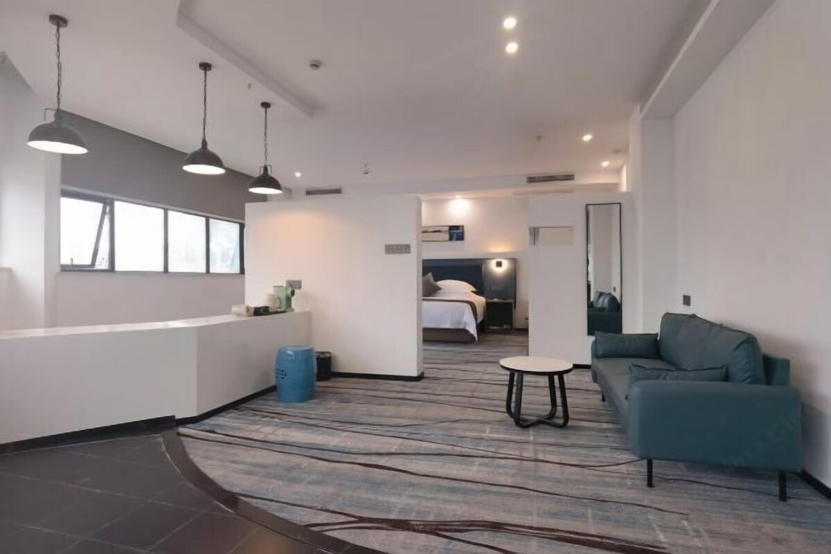 A modern hotel room with a reception desk, a seating area with a sofa and a small table, a bed, and a bathroom visible in the background.