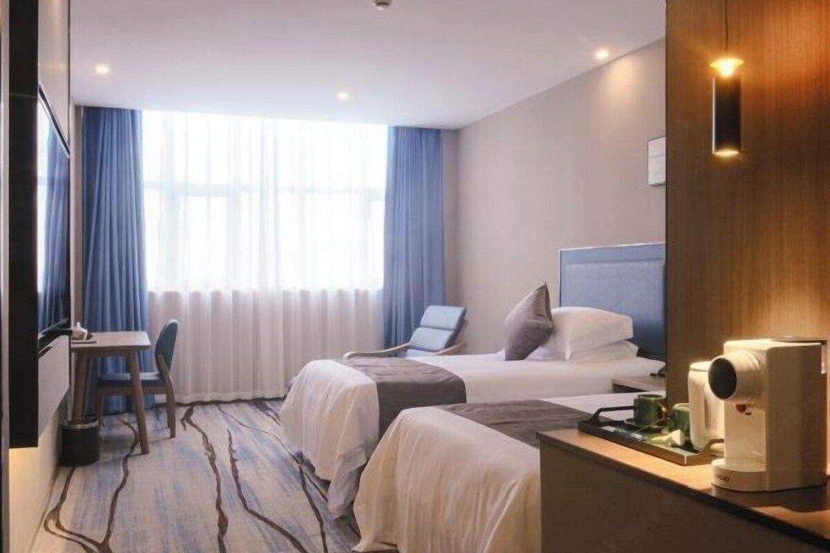 A hotel room with two beds, a desk, and a chair. There is a window with curtains, a radiator, and a wall-mounted light fixture.