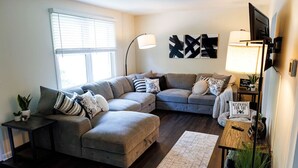 A living room with a sectional sofa, a coffee table, a lamp, and a sign on the wall. - The Cooper Home, Dogs Welcome! (Ephrata)
