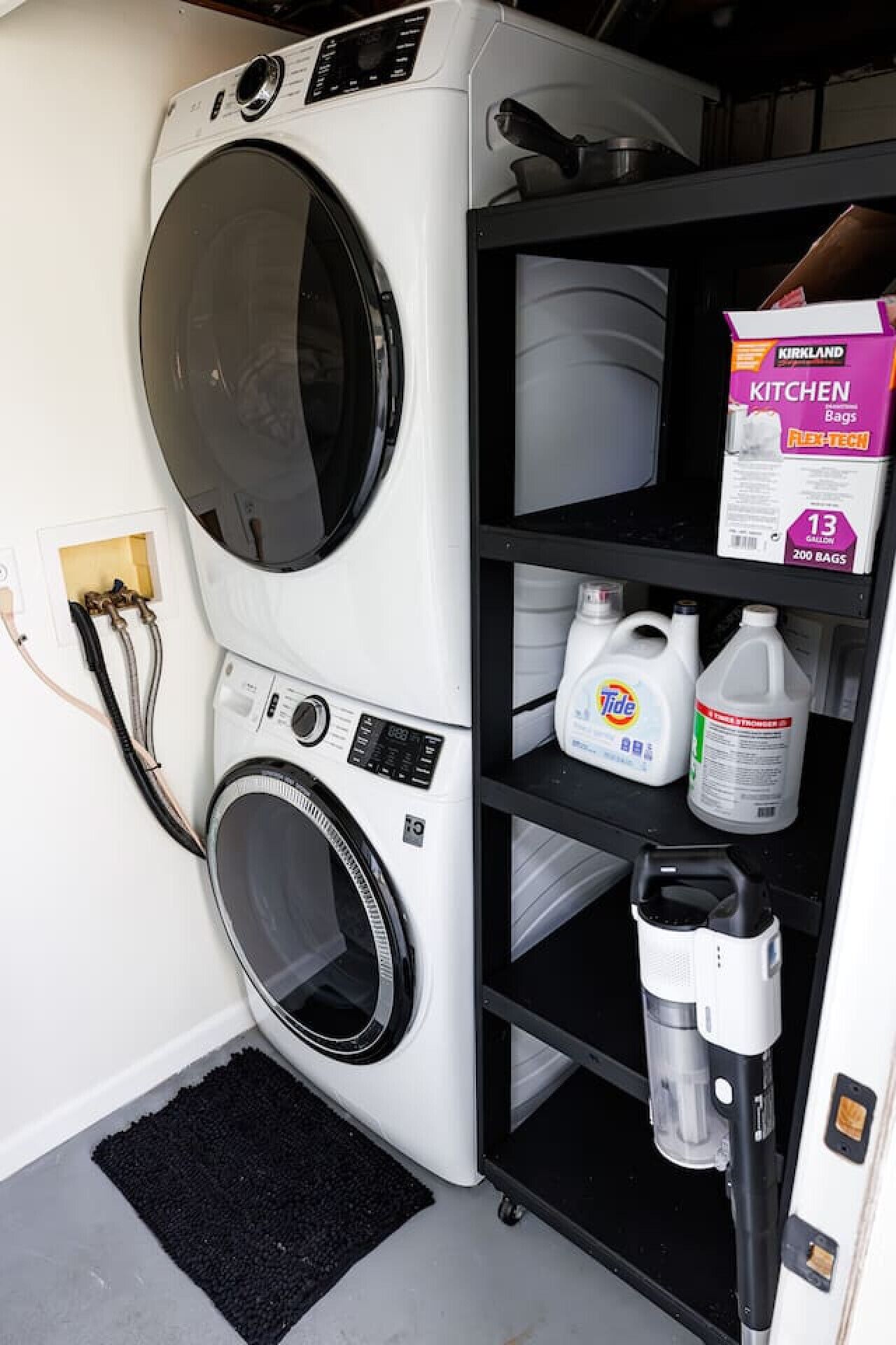 A compact laundry room with a front-loading washer and dryer, a shelf with cleaning supplies, and a black mat.