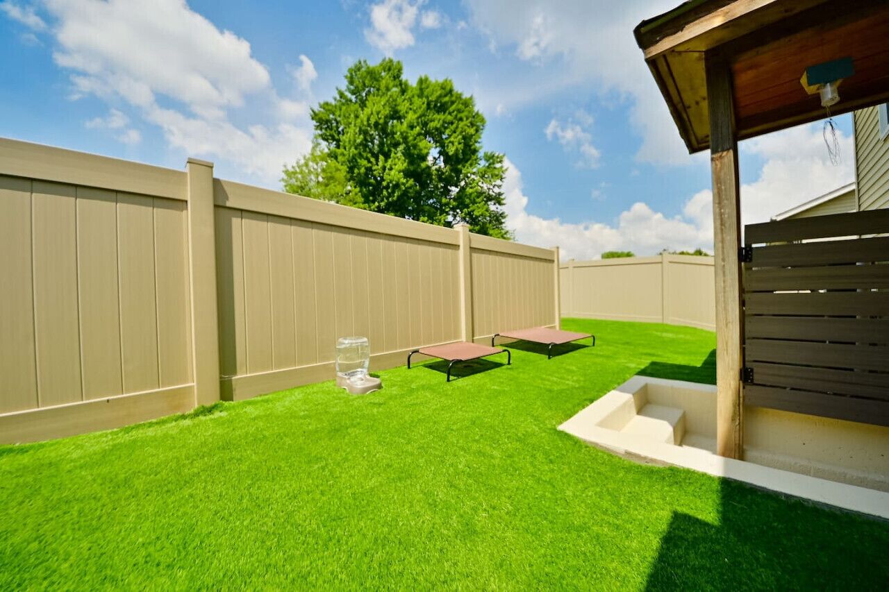 Un jardin avec une pelouse artificielle, une terrasse en bois et une clôture de couleur beige.