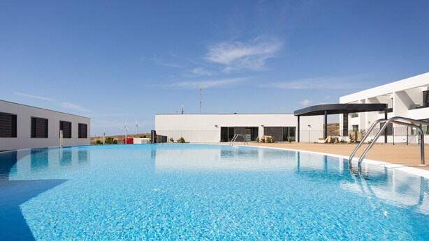 A modern pool area with clear blue water, a covered seating area, and a building with large windows and a flat roof.
