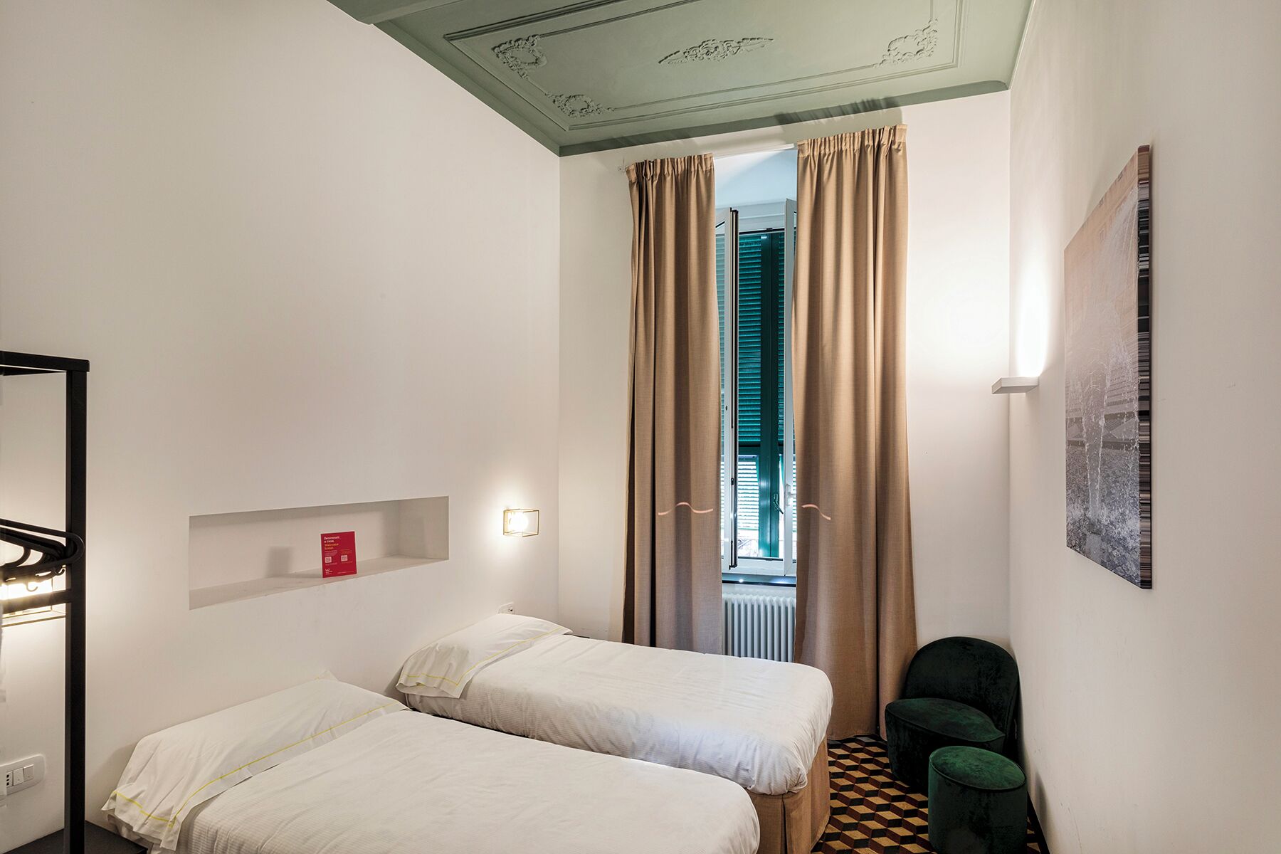 Ein Hotelzimmer mit zwei Betten, einer kleinen Ablage mit einem roten Gegenstand, einer Wandleuchte, einem gerahmten Bild und einem Schachbrettmuster auf dem Boden.