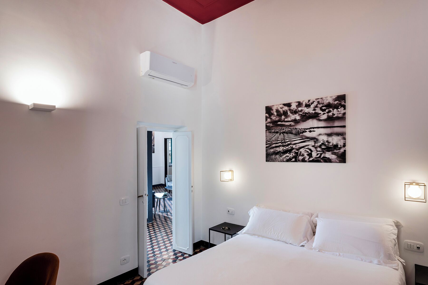 A hotel room with a bed, a door leading to a dining area, an air conditioning unit, and a wall-mounted artwork.