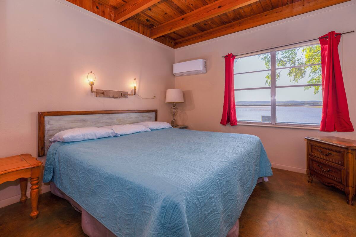 Un dormitorio con techo de madera, una cama con colcha azul, una mesita de noche, una lámpara y una ventana con cortinas rojas.