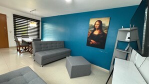 Un salon avec un mur d’accent bleu, un canapé gris, un pouf gris, une table à manger avec des chaises et un ventilateur de plafond.