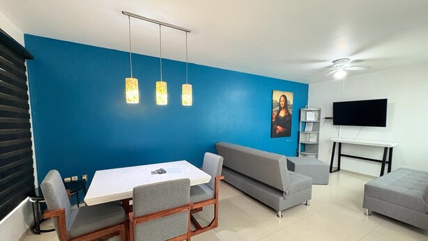 Un salon moderne avec un mur d’accent bleu, un coin repas avec une table blanche et des chaises en bois, un canapé gris, un fauteuil gris, un téléviseur fixé au mur et un ventilateur de plafond.