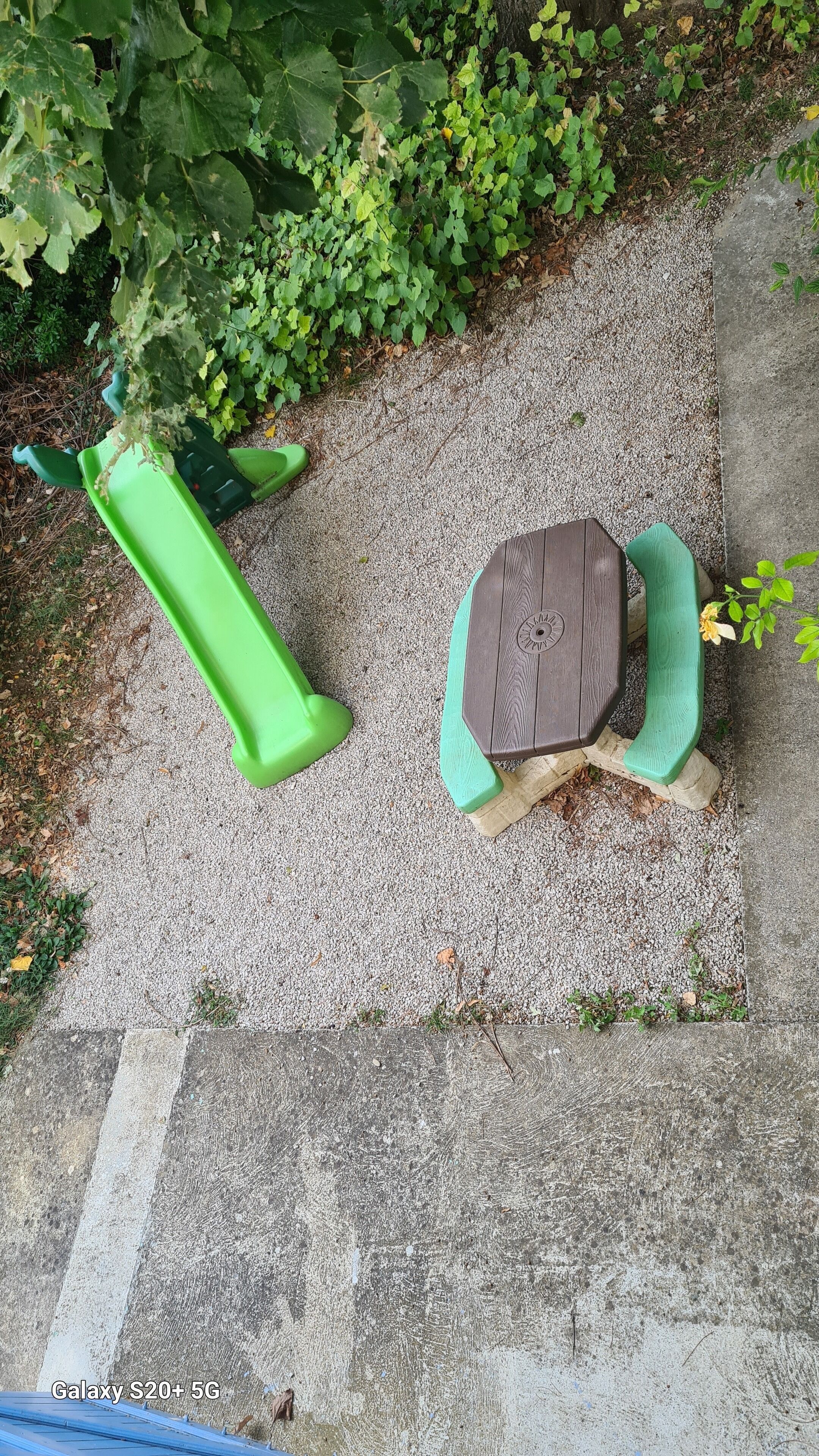 Un jardín con un sendero de hormigón, una jardinera verde y una mesa de madera con un banco.