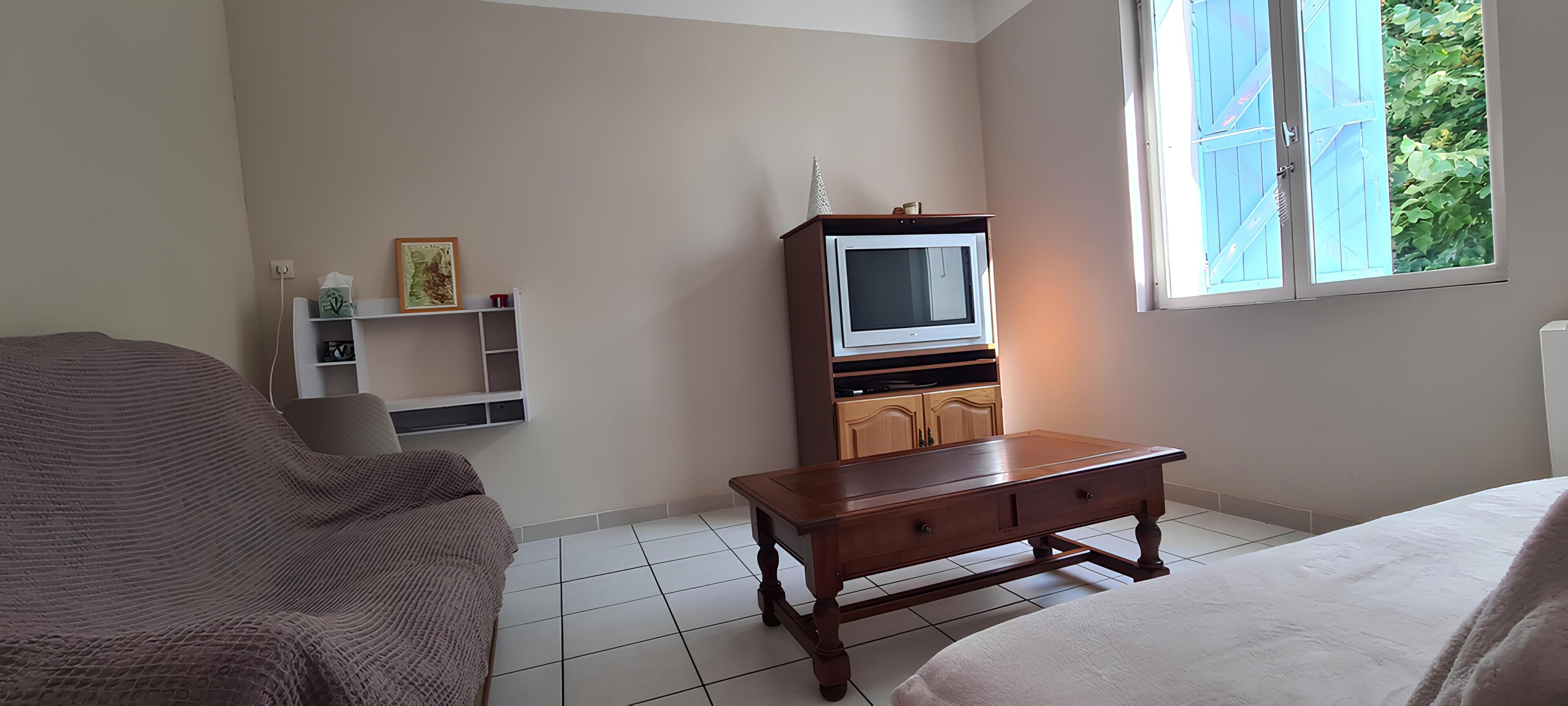 A bedroom with a bed, a wooden dresser, a television, and a window with a view of greenery.