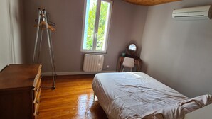 A bedroom with a wooden dresser, a bed with a white bedspread, a radiator, a window with a view of greenery, and an air conditioning unit. - Maison Avec Jardin Clôt (Montélimar)