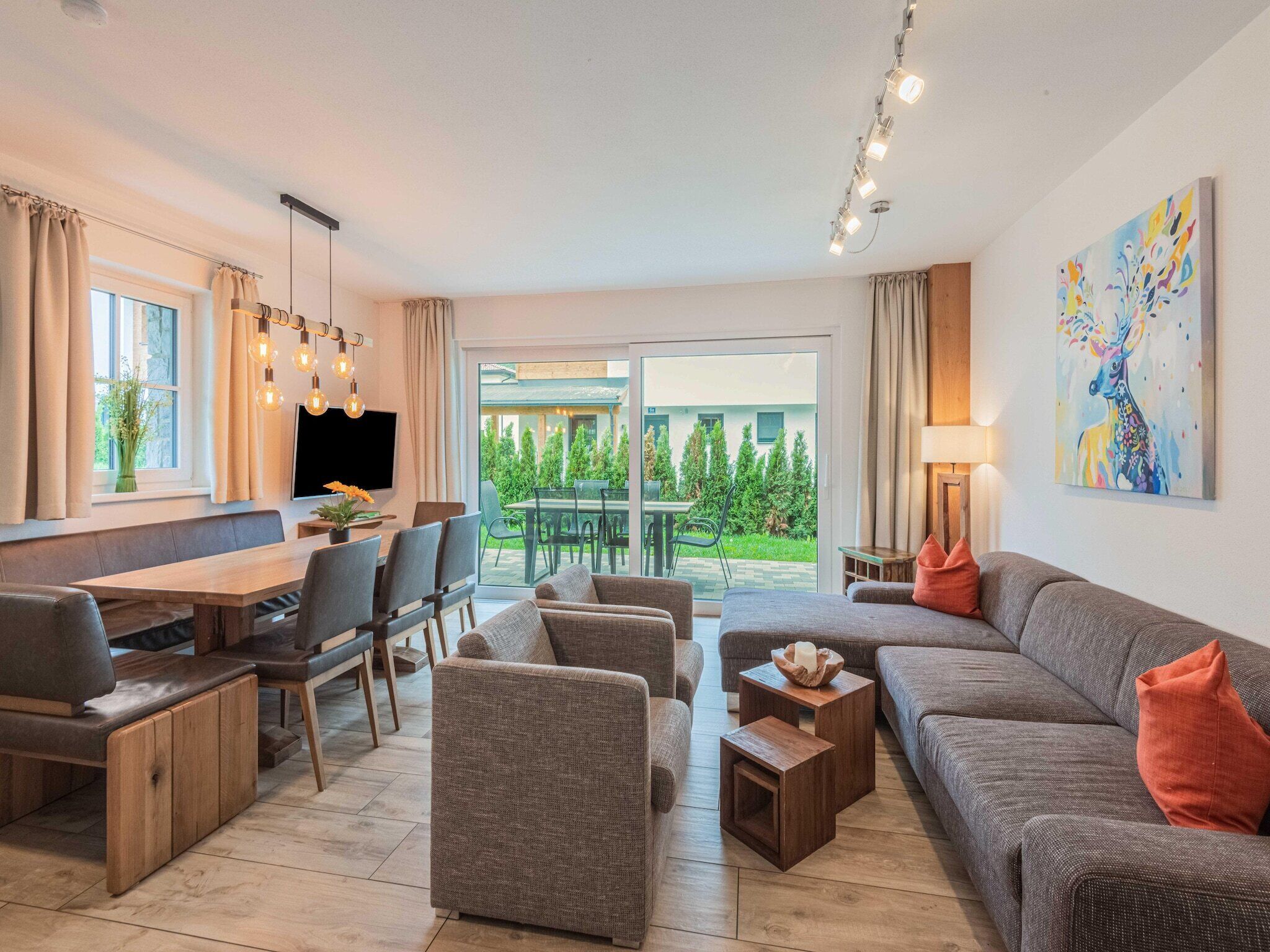 A modern living room with a sofa, chairs, a dining table, and a TV. There is a sliding glass door leading to an outdoor area with greenery.