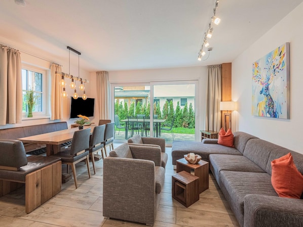A modern living room with a sofa, chairs, a dining table, and a TV. There is a sliding glass door leading to an outdoor area with greenery.