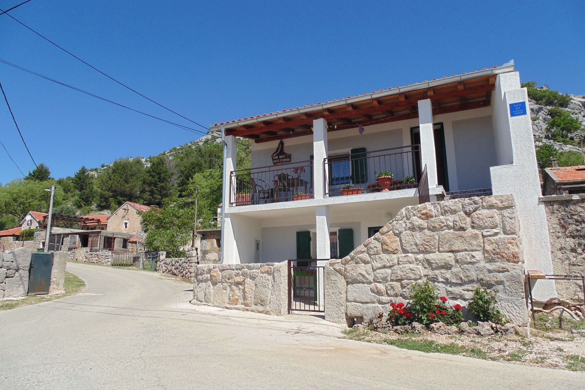 A two-story building with a stone facade, a balcony with potted plants, and a clear sky.