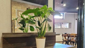 Un comptoir d’accueil agrémenté d’une plante en pot, un sol carrelé et, au fond, un espace repas.