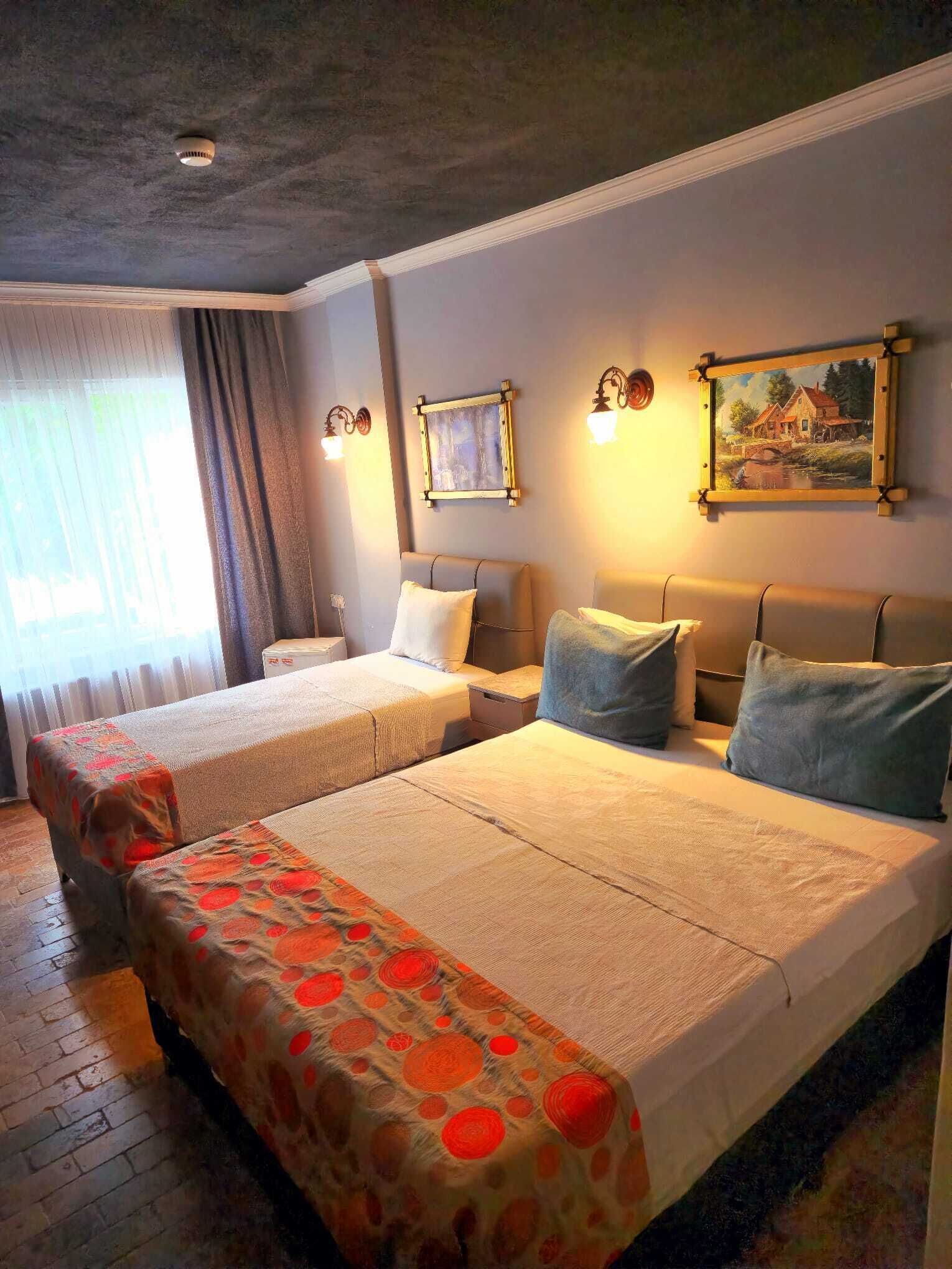 Habitación de hotel con dos camas, un ventanal con cortinas, un cuadro en la pared y una lámpara en el techo.