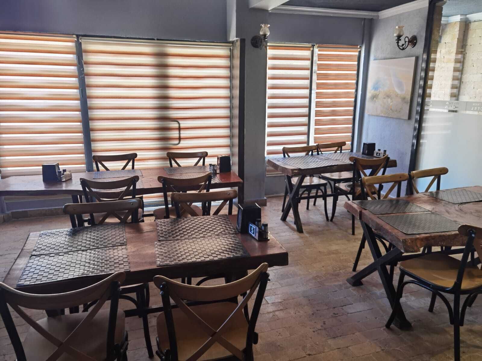 L’intérieur d’un restaurant avec des tables et des chaises en bois, de grandes fenêtres équipées de stores horizontaux et un mur de briques agrémenté d’un élément décoratif.