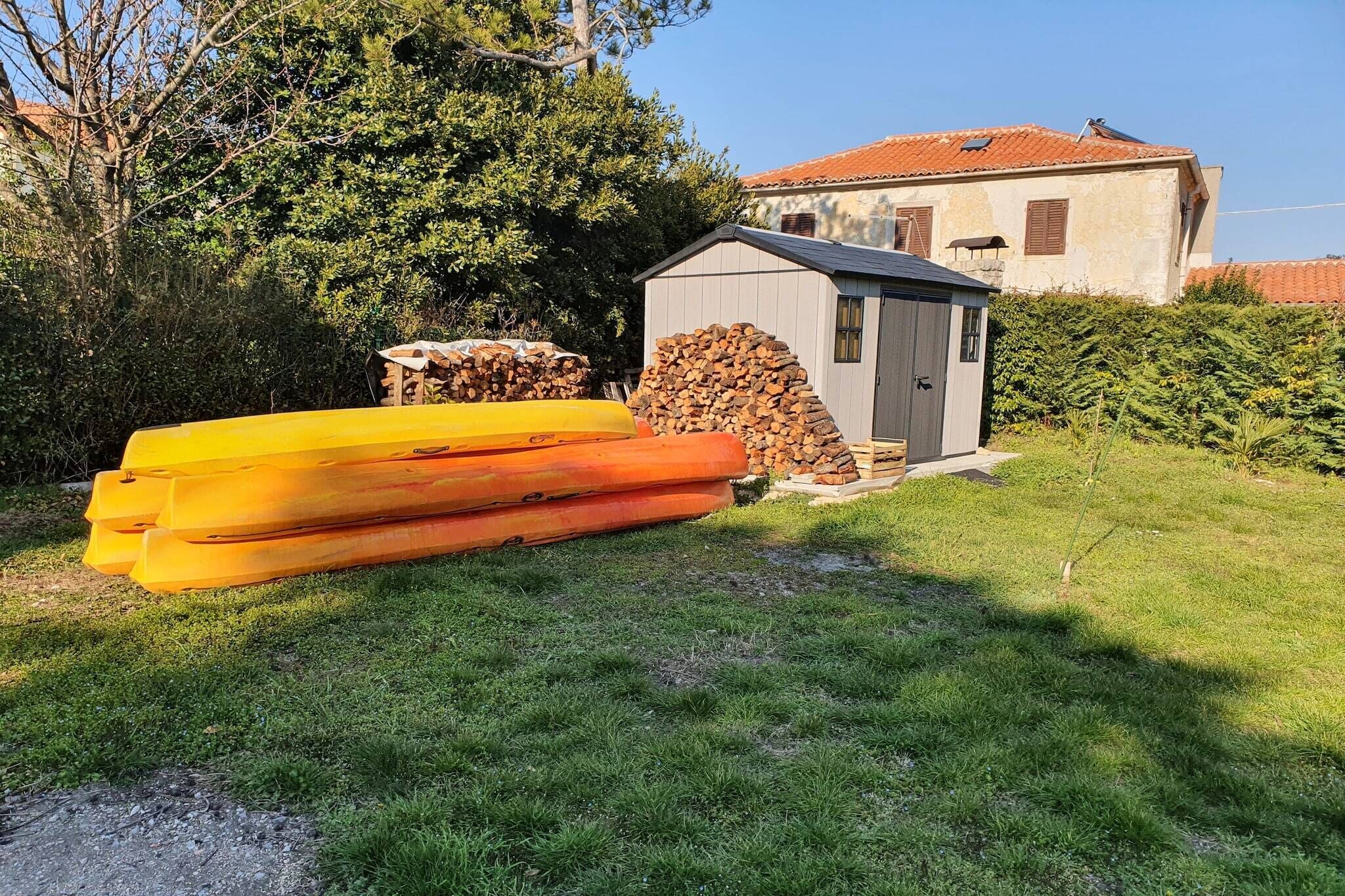 Un área cubierta de césped con una pila de kayaks amarillos y un montón de leña junto a un cobertizo pequeño.