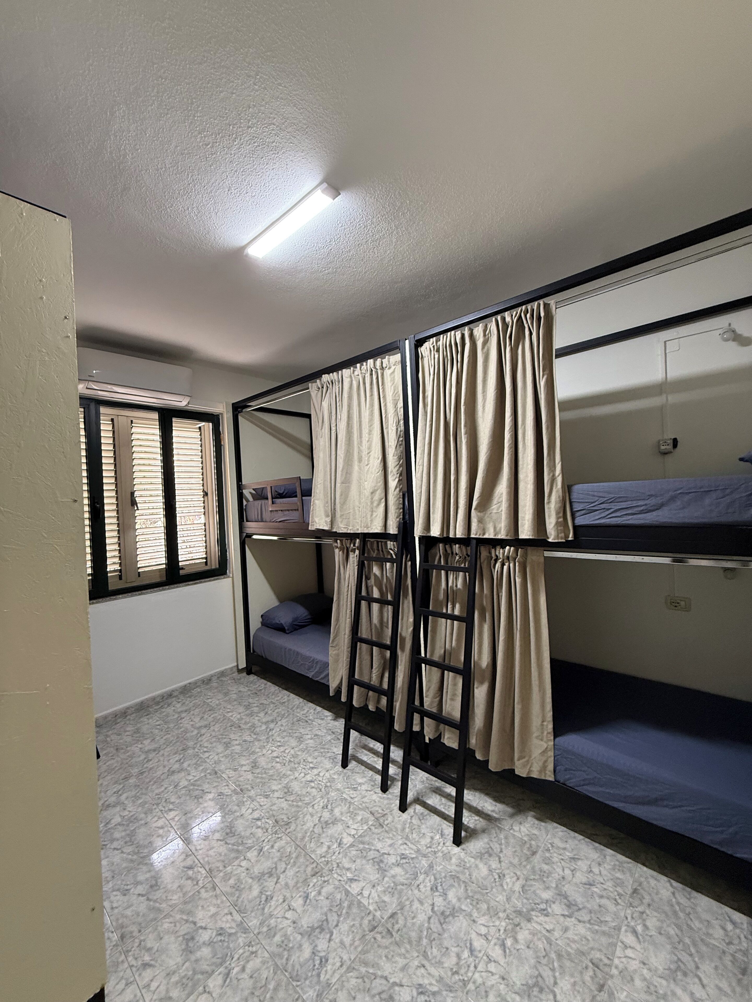 A dormitory room with bunk beds, a window with blinds, and an air conditioning unit.