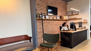 A modern reception area with a brick wall, a flat-screen TV, and a wooden counter with a laptop and a coffee machine. - Shkodra Hostel (Shkodër)