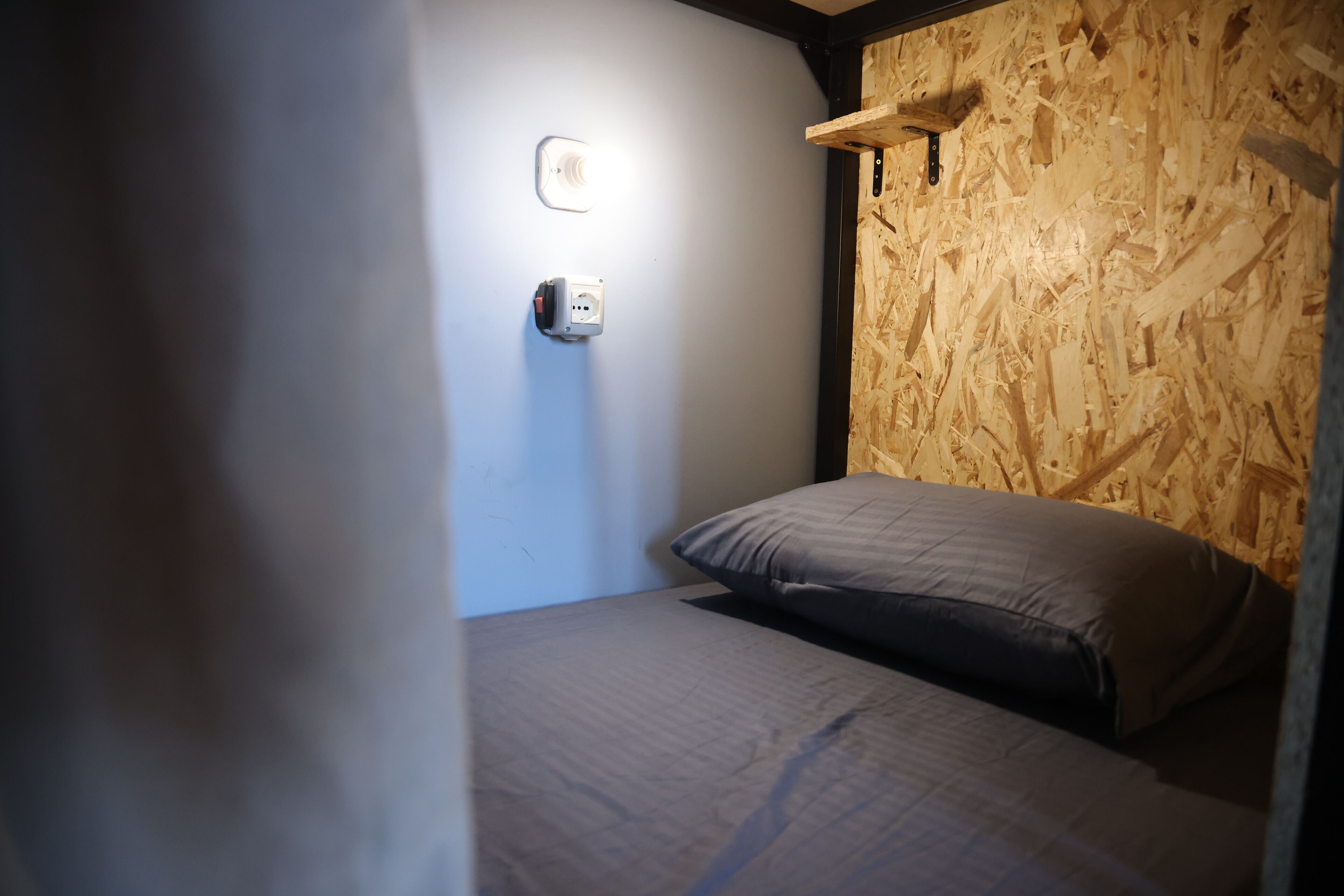A small, dimly lit room with a single bed, a wooden wall, and a wall socket.