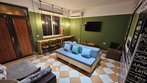 A cozy living room with a sofa, a dining table, and chairs. There is a window with blinds, a flat-screen TV, and a decorative wall with city names. - Shkodra Hostel (Shkodër)