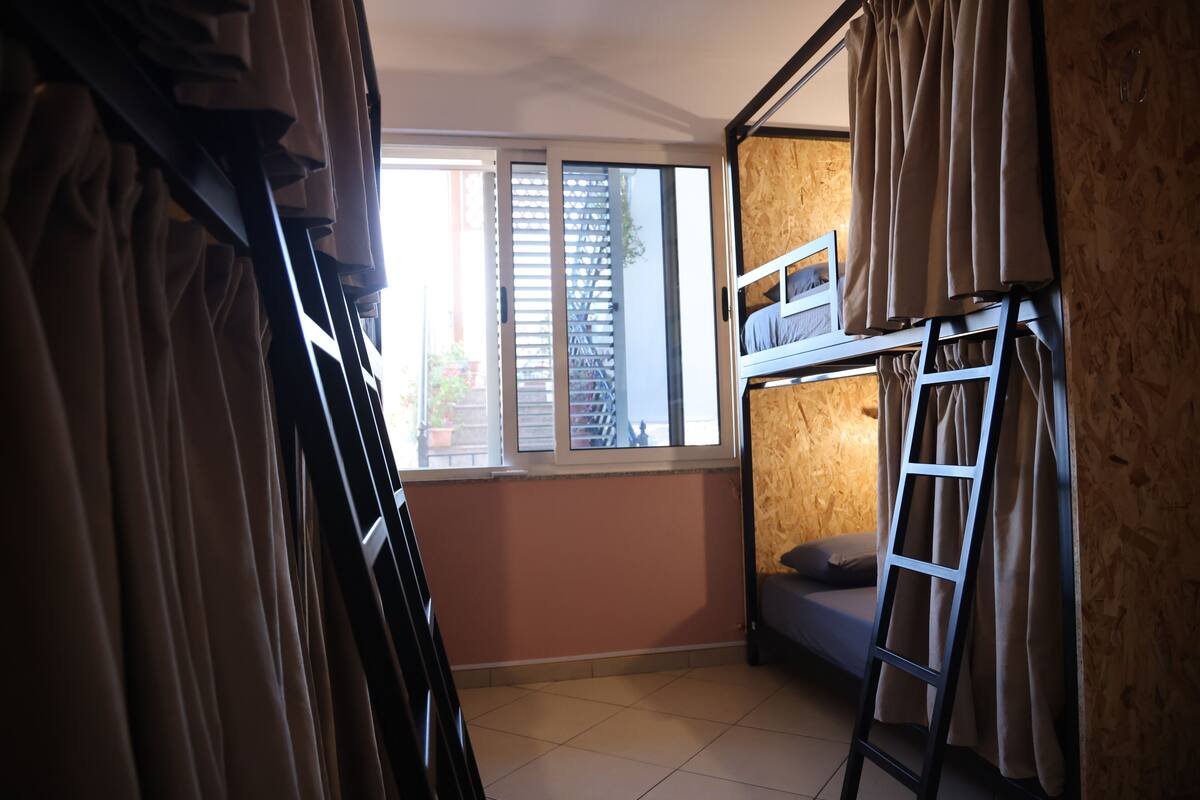 Une chambre avec un lit superposé, une fenêtre avec des stores, une échelle et une vue sur l’extérieur.